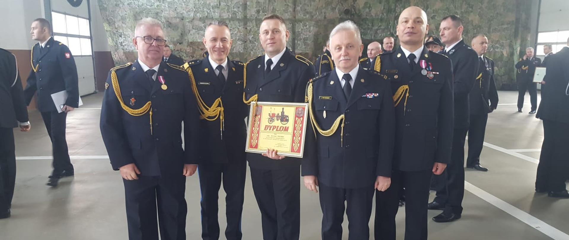 Zdjęcie wykonane na ternie Komendy Powiatowej Państwowej Straży Pożarnej w Ciechanowie. Na zdjęciu widać na pierwszym panie stojących obok siebie pięciu funkcjonariuszy. Funkcjonariusze ubrani są w umundurowanie galowe w kolorze ciemnym granatowym. Wśród nich znajdują się komendant główny oraz mazowiecki komendant wojewódzki Państwowej Straży Pożarnej. Dwóch funkcjonariuszy do munduru przypięte ma medale a jeden z nich w rękach trzyma pamiątkowy dyplom, które to otrzymali podczas apelu. W oddali za ich plecami widać innych strażaków również umundurowanych w umundurowanie galowe. 
