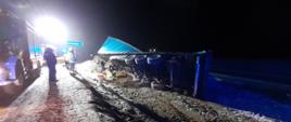 Na zdjęciu widać pobocze drogi oraz przewróconą na prawą stronę naczepę samochodu ciężarowego. W lewej części zdjęcia stoi samochód pożarniczy. Widać także strażaków prowadzących działania ratownicze.