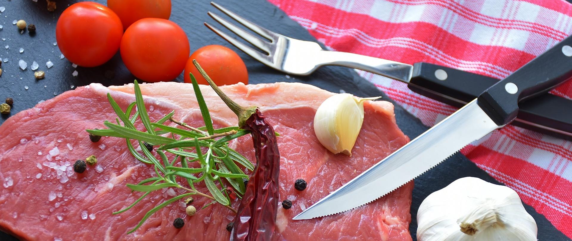 Na desce znajduje się surowy stek posypany ziarnami pieprzu i solą, na którym leży ząbek czosnku i ostra papryczka. Na desce obok steku jest główka czosnku, pomidorki koktajlowe, nóż i widelec.