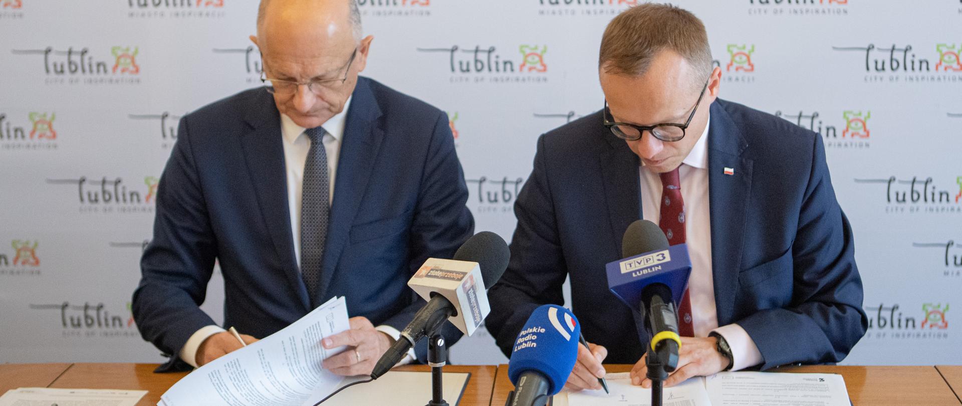 na stole leżą teksty umowy, które podpisują przedstawiciele miasta i ministerstwa, widać także mikrofonu postawione na stole