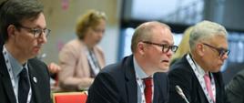Do mikrofonu przemawia mężczyzna siedzący przy stole konferencyjnym między dwoma innymi mężczyznami