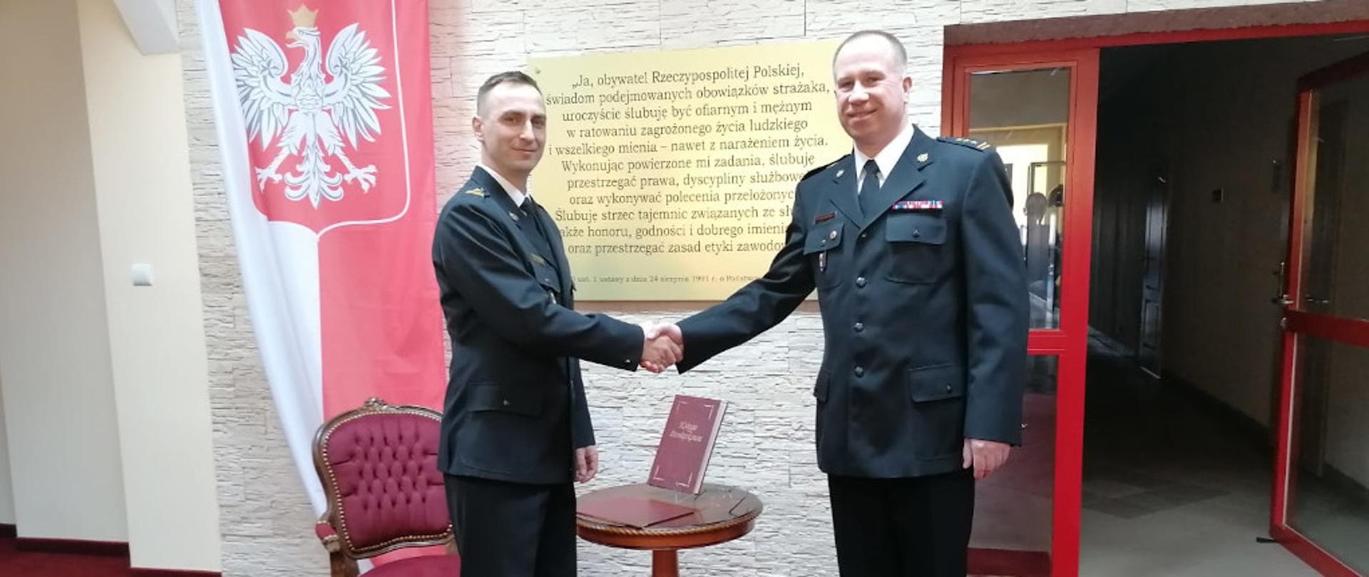 Na zdjęciu znajdują się dwaj strażacy w umundurowaniu galowym podający sobie prawe dłonie. Strażacy umiejscowieni są w pomieszczeniu sali tradycji Komendy Powiatowej Państwowej Straży Pożarnej w Jędrzejowie. W tle widnieją na ścianie słowa roty ślubowania strażaków oraz biało- czerwona flaga.