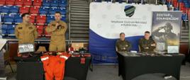Zdjęcie panoramiczne zrobione wewnątrz dużej hali sportowej podczas targów pracy. Na pierwszym planie, po lewej, stoją dwaj mężczyźni w piaskowych mundurach strażackich; jeden z nich przemawia do mikrofonu. Przed nimi znajdują się dwa stoły przykryte czarnym obrusem, na których wyeksponowano sprzęt ratowniczy: jaskrawo pomarańczowy kombinezon, czerwoną apteczkę modułową z napisem „URAZOWY”, otwartą czarną walizkę ze sprzętem medycznym oraz fantom szkoleniowy. Za stołami, po prawej stronie, siedzą dwaj inni mężczyźni w wojskowych mundurach polowych. Tło stanowi ścianka reklamowa z logo i napisem „Wojskowe Centrum Rekrutacji w Kołobrzegu” oraz roll-up z hasłem „ZOSTAŃ ŻOŁNIERZEM”. W głębi widać puste trybuny hali z czerwonymi i niebieskimi krzesełkami oraz oznaczenia sektorów „C” i „D”.