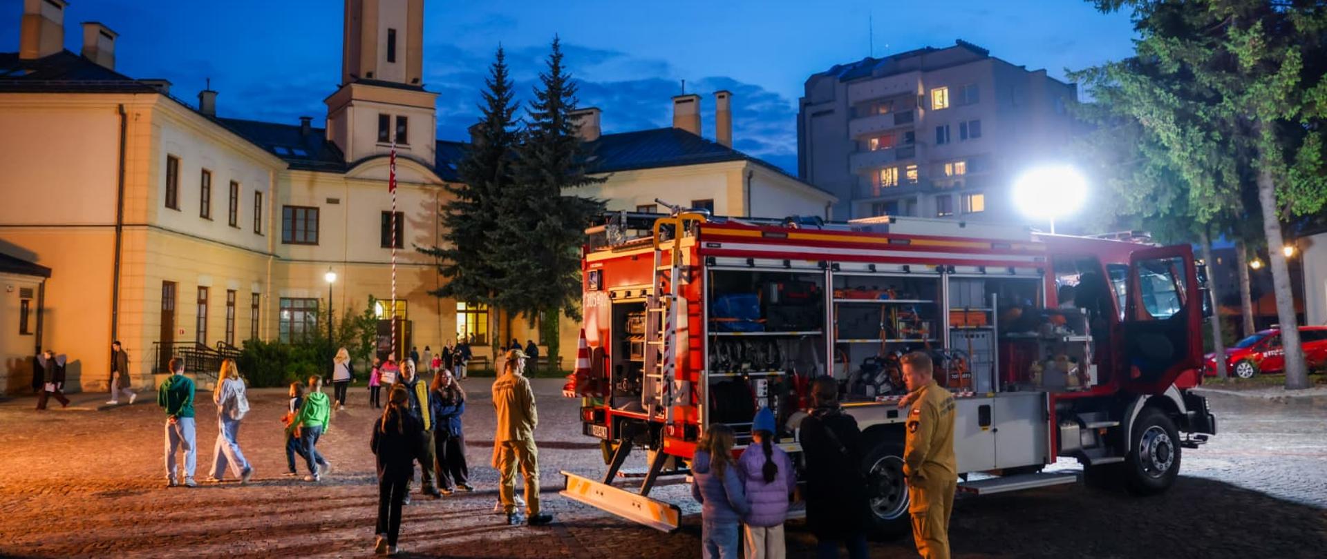 Zdjęcie przedstawia wóz strażacki z otwartymi skrytkami sprzętowymi zaparkowany wieczorem na miejskim placu. Wokół pojazdu stoją strażacy oraz zwiedzający, w tle widoczna jest zabytkowa wieża strażacka.