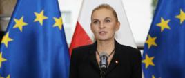 Minister Nowacka stoi i mówi do mikrofonu na stojaku, za nią pod ścianą rząd flag Polski i UE.