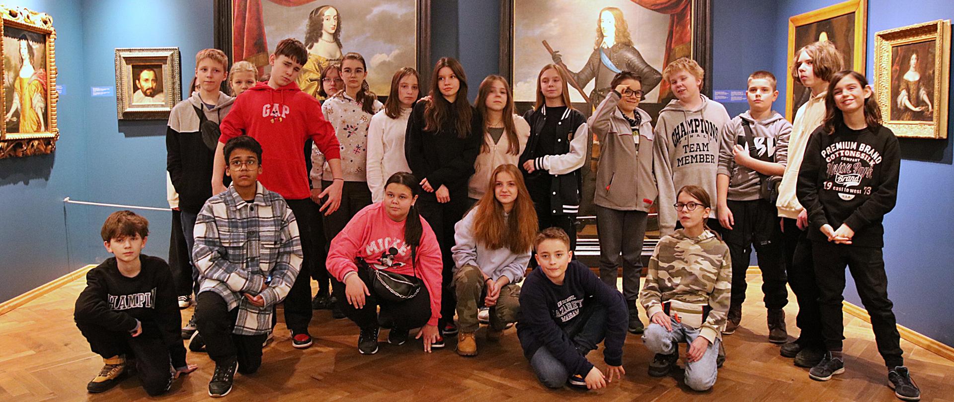 Grupa młodzieży stoi w pomieszczeniu, na niebieskich ścianach wiszą obrazy.