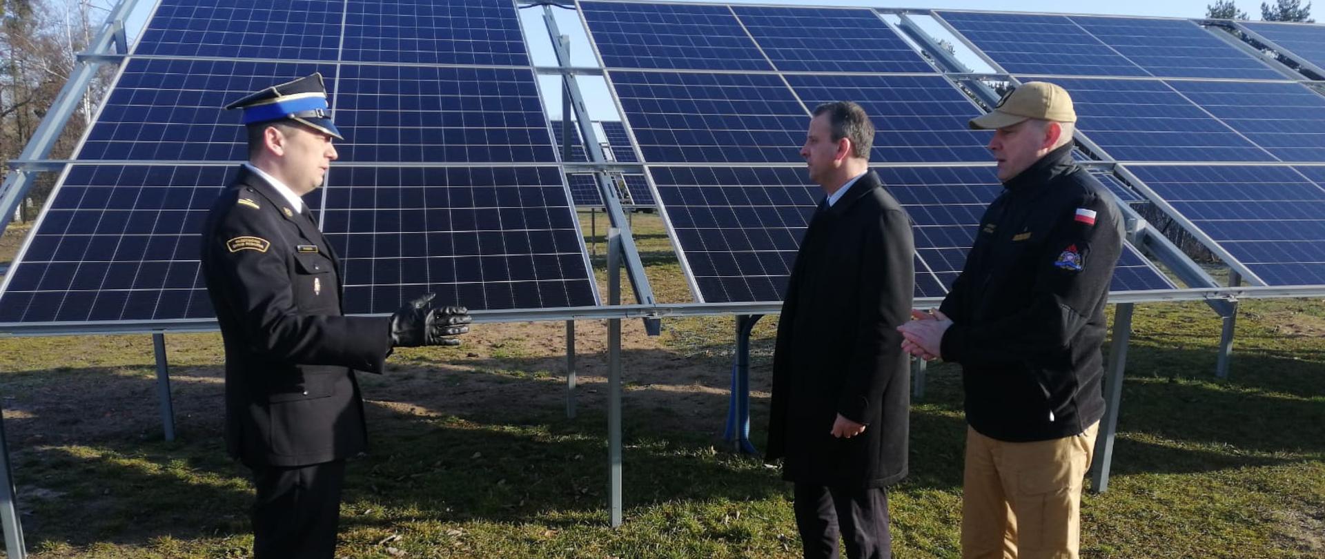 dwóch strażaków i mężczyzna w płaszczu stoją na trawiastej nawierzchni przed panelami fotowoltaicznymi