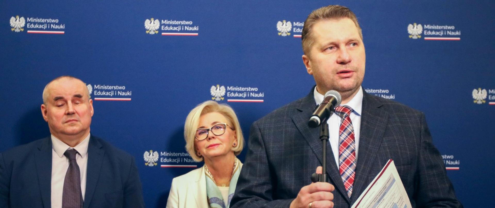 Minister Czarnek stoi i mówi do mikrofonu, za nim stoi wiceminister Machałek w białym ubraniu i mężczyzna w szarym garniturze, za nimi granatowa ściana.