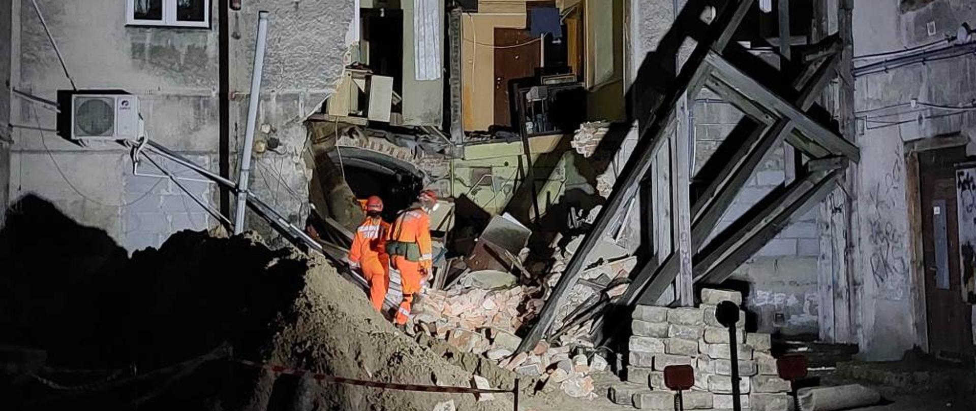 Na zdjęciu widać budynek mieszkalny – kamienicę z częściowo zawaloną ścianą. Na gruzowisku znajduje się pies poszukiwawczy wraz z dwojgiem strażaków ze specjalnej grupy poszukiwawczo ratowniczej. Ratownicy ubrani są w pomarańczowe kombinezony. Na głowach mają kaski wyposażone w latarki. Z uwagi na porę nocną teren akcji jest oświetlony. 