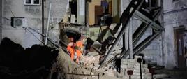 Na zdjęciu widać budynek mieszkalny – kamienicę z częściowo zawaloną ścianą. Na gruzowisku znajduje się pies poszukiwawczy wraz z dwojgiem strażaków ze specjalnej grupy poszukiwawczo ratowniczej. Ratownicy ubrani są w pomarańczowe kombinezony. Na głowach mają kaski wyposażone w latarki. Z uwagi na porę nocną teren akcji jest oświetlony. 
