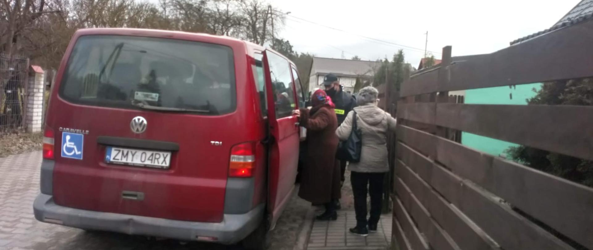 Na zdjęciu widzimy strażaka ubranego w umundurowanie koszarowe, pomagającego seniorce wejść do samochodu typu bus.