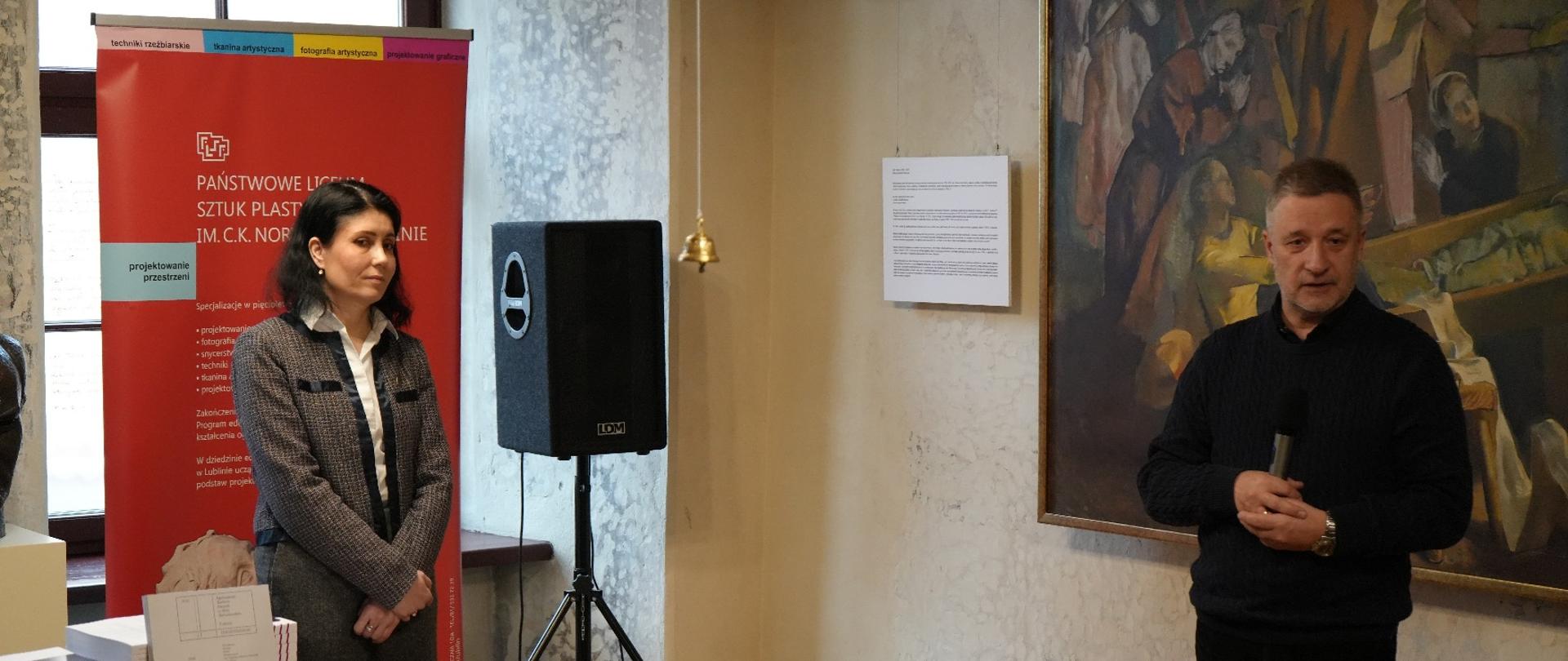 Zdjęcie w pomieszczeniu w Muzeum Józefa Czechowicza w Lublinie. Dwie osoby stoją przed czerwonym banerem z białym napisem „PAŃSTWOWE LICEUM SZTUK PLASTYCZNYCH IM. C.K. NORWIDA W LUBLINIE” oraz dużą reprodukcją obrazu na ścianie. Po lewej stronie stoi kobieta w szaro-czarnym żakiecie i ciemnych spodniach, ze skrzyżowanymi rękami. Po prawej stronie stoi mężczyzna w czarnym swetrze i spodniach, trzymający mikrofon i przemawiający. Pomiędzy nimi znajduje się duży, czarny głośnik LDM na statywie. Na stole po lewej leżą dokumenty lub książki, a na dole zdjęcia widoczna jest część publiczności słuchającej prelegentów.