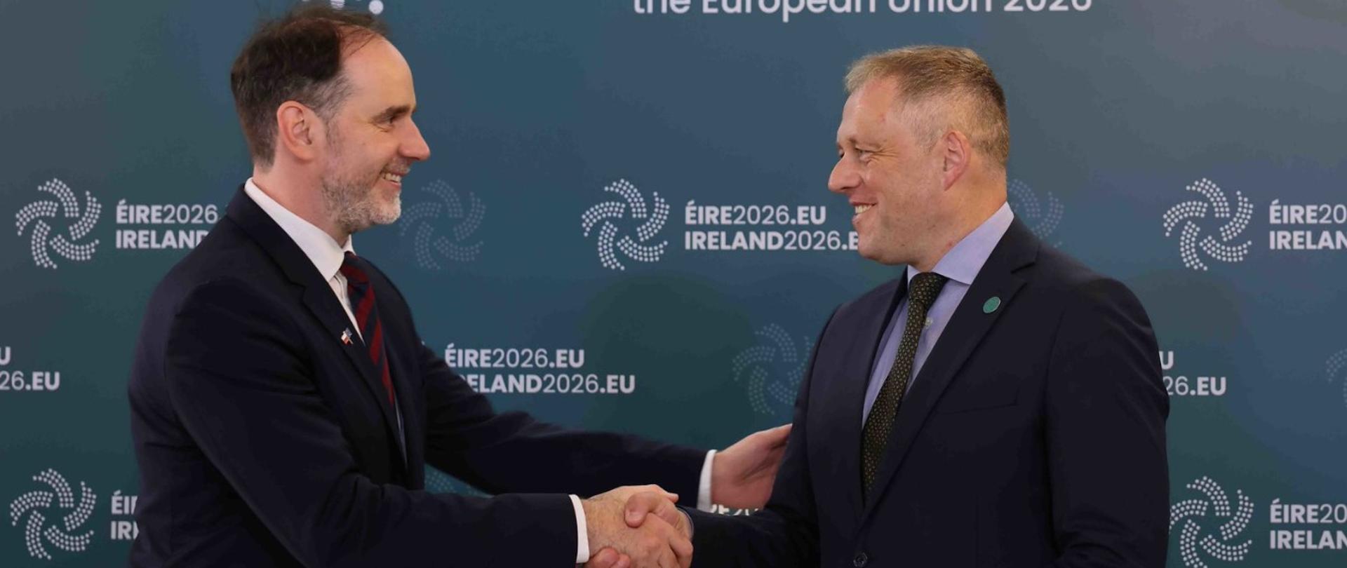 Dwóch mężczyzn w garniturach podaje sobie dłonie podczas spotkania. W tle znajduje się ścianka z logo irlandzkiej prezydencji w Radzie Unii Europejskiej 2026 oraz napisem „EIRE2026.EU / IRELAND2026.EU”. Mężczyźni uśmiechają się do siebie.
