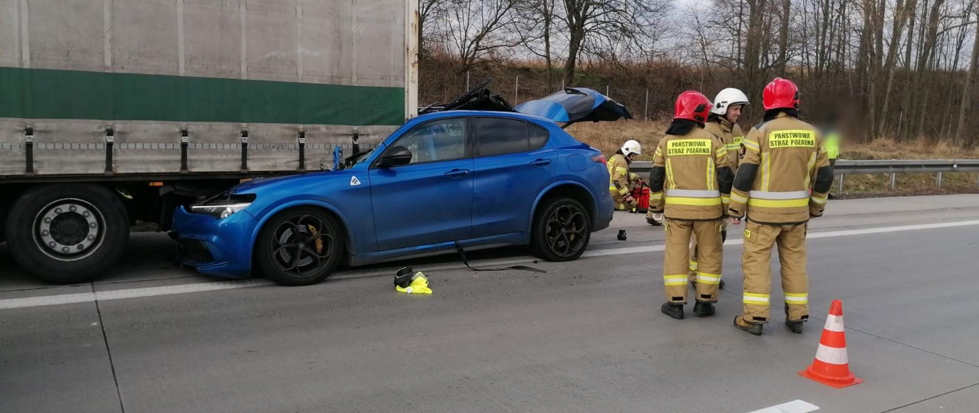 Obraz przedstawia samochód osobowy wbity pod naczepę samochodu ciężarowego. Przy pojeździe strażacy w umundurowaniu specjalnym. Pora dzienna.