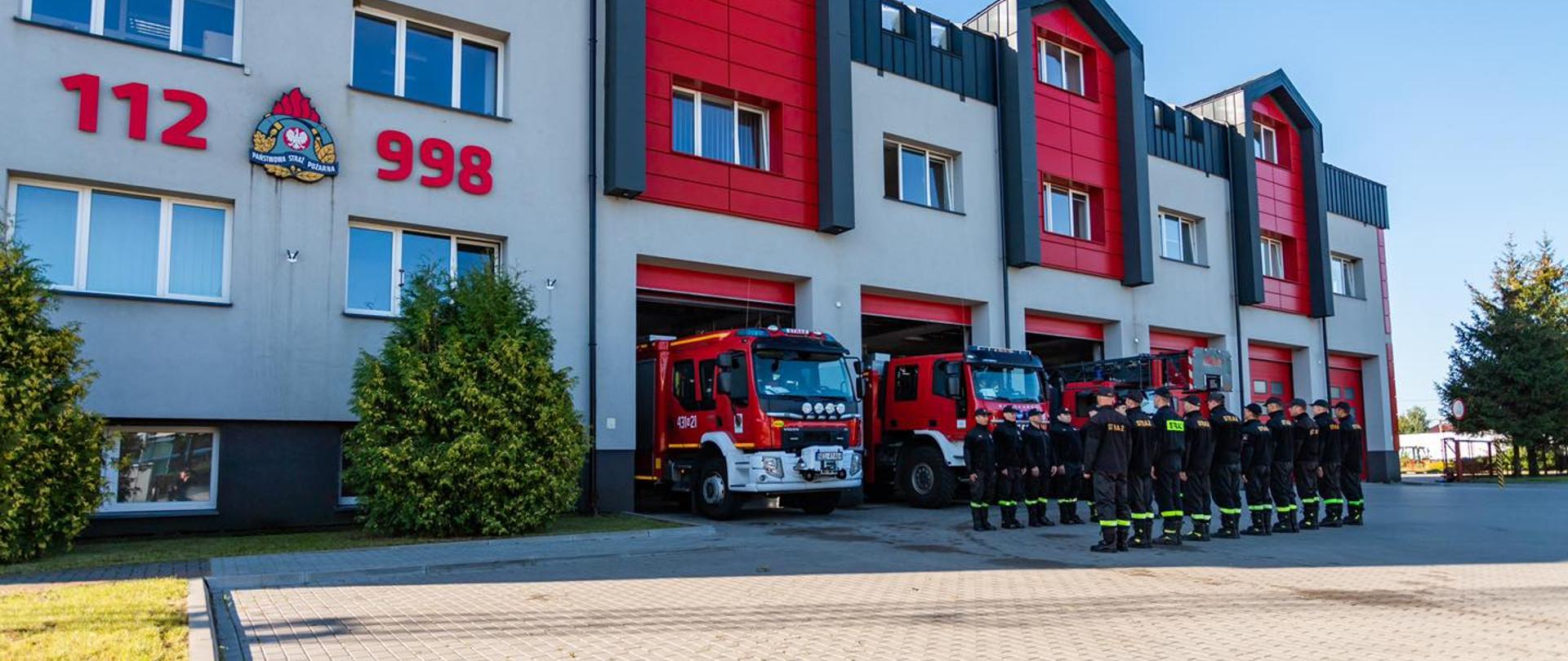 Na zdjęciu strażacy podczas zmiany służby oddający hołd zmarłemu tragicznie koledze. W tle budynek komendy powiatowej i pojazdy pożarnicze.