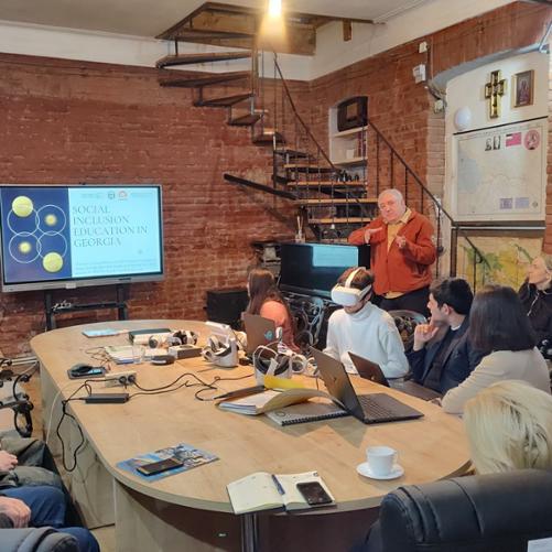 Przy owalnym stole ustawionym obok telewizora, na którym jest wyświetlana prezentacja, siedzi grupa osób, w tym mężczyzna, który ma założone okulary VR