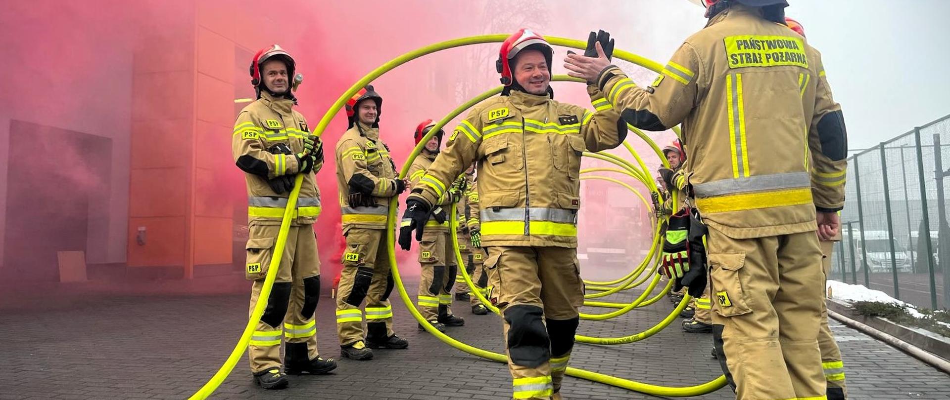 Na zdjęciu widzimy uroczysty apel strażaków z Państwowej Straży Pożarnej (PSP). Strażacy stoją w dwóch rzędach, tworząc symetryczną formację, a jeden z nich oddaje salut przed przełożonym w granatowym mundurze.
W tle znajduje się wóz strażacki z numerem operacyjnym oraz siatkowe ogrodzenie z boiskiem. Atmosfera na zdjęciu jest oficjalna, a pogoda wydaje się mglista i chłodna, co potęguje surowy klimat wydarzenia.
