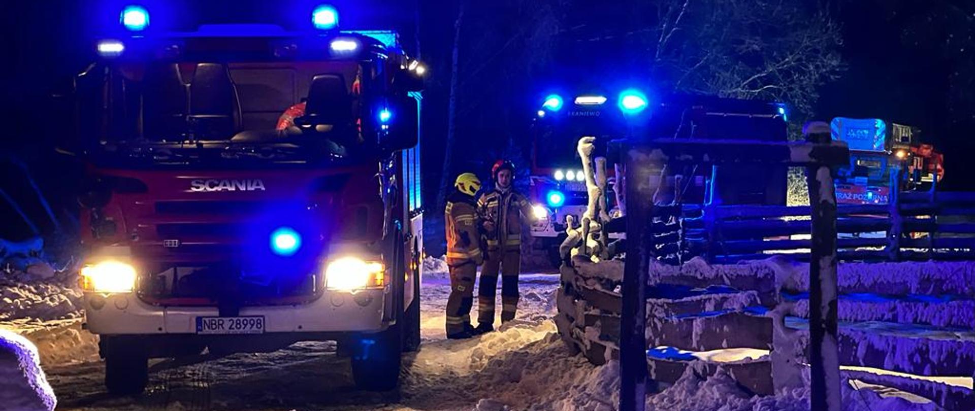 Zdjęcie zrobione nocą. Na na zaśnieżonej drodze stoją trzy samochody strażackie na światłach i maja włączone niebieskie świtała ostrzegawcze. Przy pierwszym samochodzie stoi dwóch strażaków.