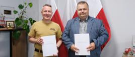 Dwaj mężczyźni na tle flag narodowych. Trzymają w rękach teczki z podpisaną wcześniej umową