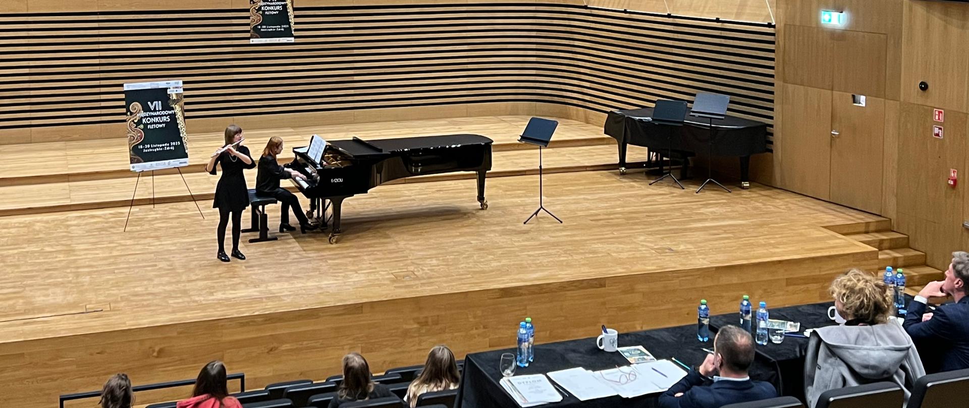 Dziewczyna gra na flecie, kobieta akompaniuje jej na fortepianie, na widowni widać od tyłu Jury i publiczność.