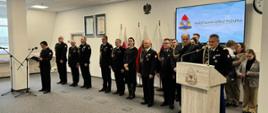 Strażak w mundurze wyjściowym ze sznurem stoi przy mównicy i przemawia za nimi stoją inni strażacy w mundurach wyjściowych ze sznurem zaproszeni goście oraz rodziny obok przed mikrofonem stoi funkcjonariusz prowadzący uroczystość. 
