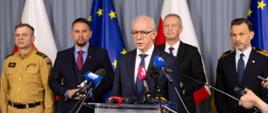 Wiceminister Wiesław Leśniakiewicz stoi przy mikrofonie. Za nim wojewoda podlaski, rzecznik ministra spraw wewnętrznych, mężczyzna w garniturze i strażak