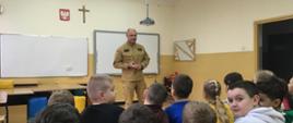 Na zdjęciu widać funkcjonariusza PSP oraz policji ubranego w ubranie służbowe w trakcie pogadanki dla uczniów szkoły
podstawowej oraz przedszkola. Znajdują się na sali.