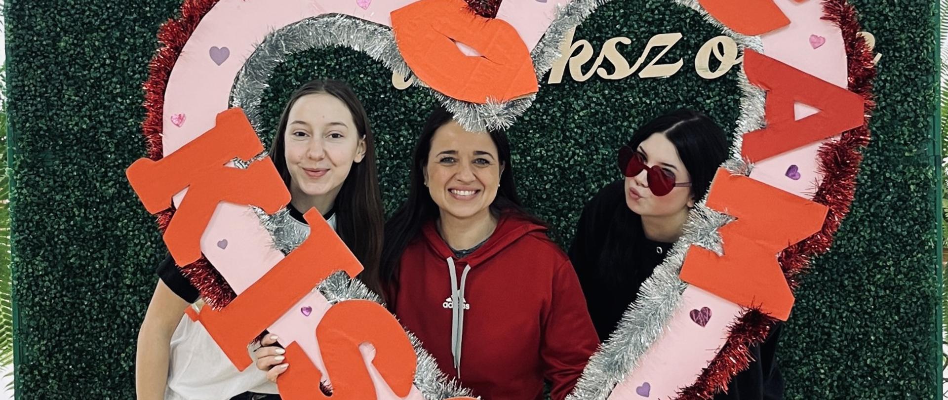 Trzy kobiety stoją przed zieloną ścianką i trzymają dużą, dekoracyjną ramkę w kształcie serca z napisem