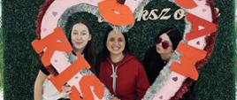 Trzy kobiety stoją przed zieloną ścianką i trzymają dużą, dekoracyjną ramkę w kształcie serca z napisem