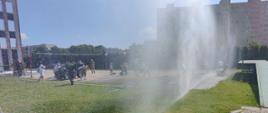 Widok kurtyn gaśniczych tryskających wodę a za nimi dzieci i strażacy na placu
