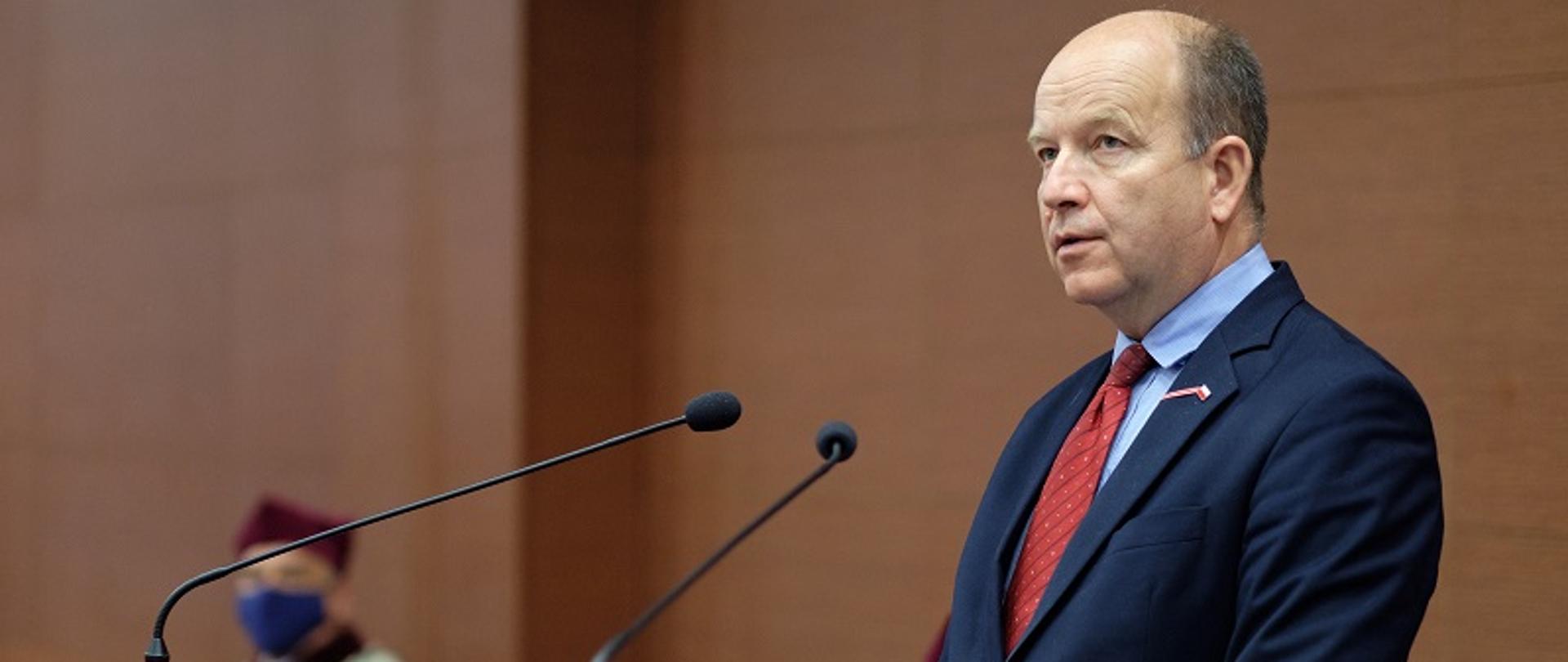 Na zdjęciu Wojewoda Mazowiecki podczas przemówienia, ubrany w granatowy garnitur i czerwony krawat. 