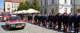 Na pierwszym planie zdjęcia widzimy zabytkowy strażacki samochód osobowy marki "POLONEZ" koloru czerwonego, który bierze udział w defiladzie strażackiej. Za samochodem strażackim widzimy kolejne zabytkowe samochody strażackie. Defiladzie przyglądają się zebrane osoby strażacy, zaproszeni goście. W oddali widzimy część budynku ratusza koloru białego oraz budynki mieszkalne ( kamienice ) koloru piaskowego. 