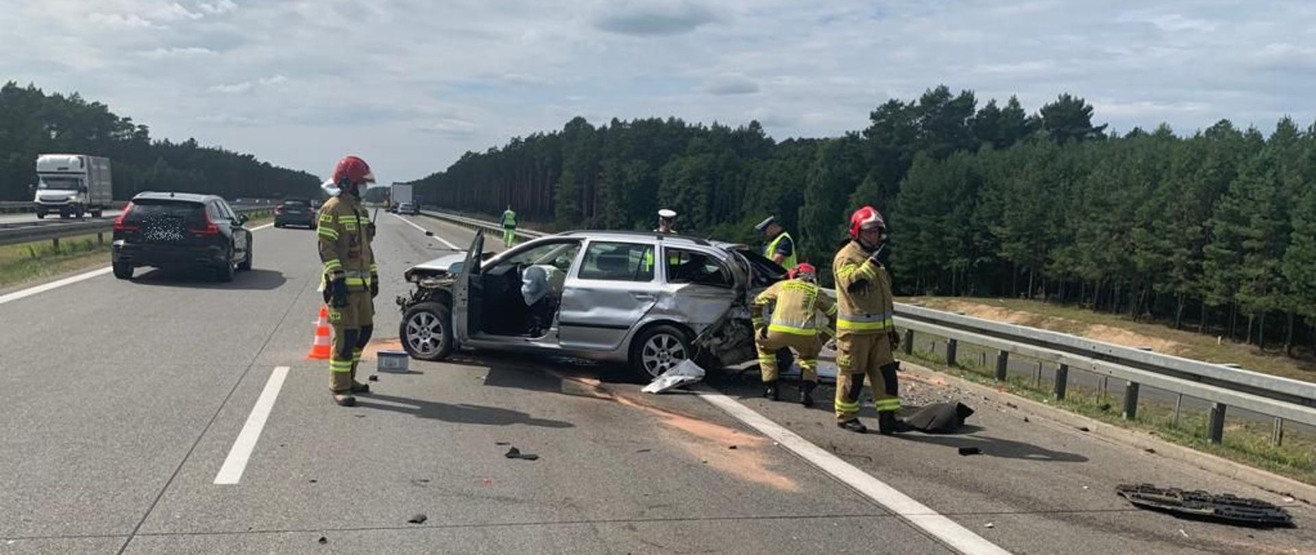 Dzień. Stojący w poprzek jezdni rozbita Skoda Octavia, przy niej strażacy.