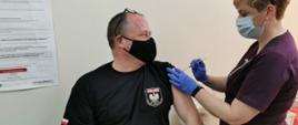 Strażak w umundurowaniu dowódczo sztabowym w czarnym podkoszulku przyjmuje szczepionkę