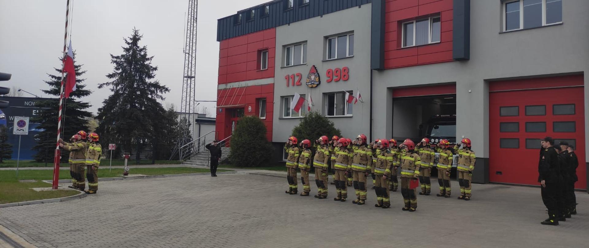 Zdjęcie przedstawia funkcjonariuszy KP PSP w Ełku w trakcie uroczystego podniesienia flagi państwowej, w tle strażnica.