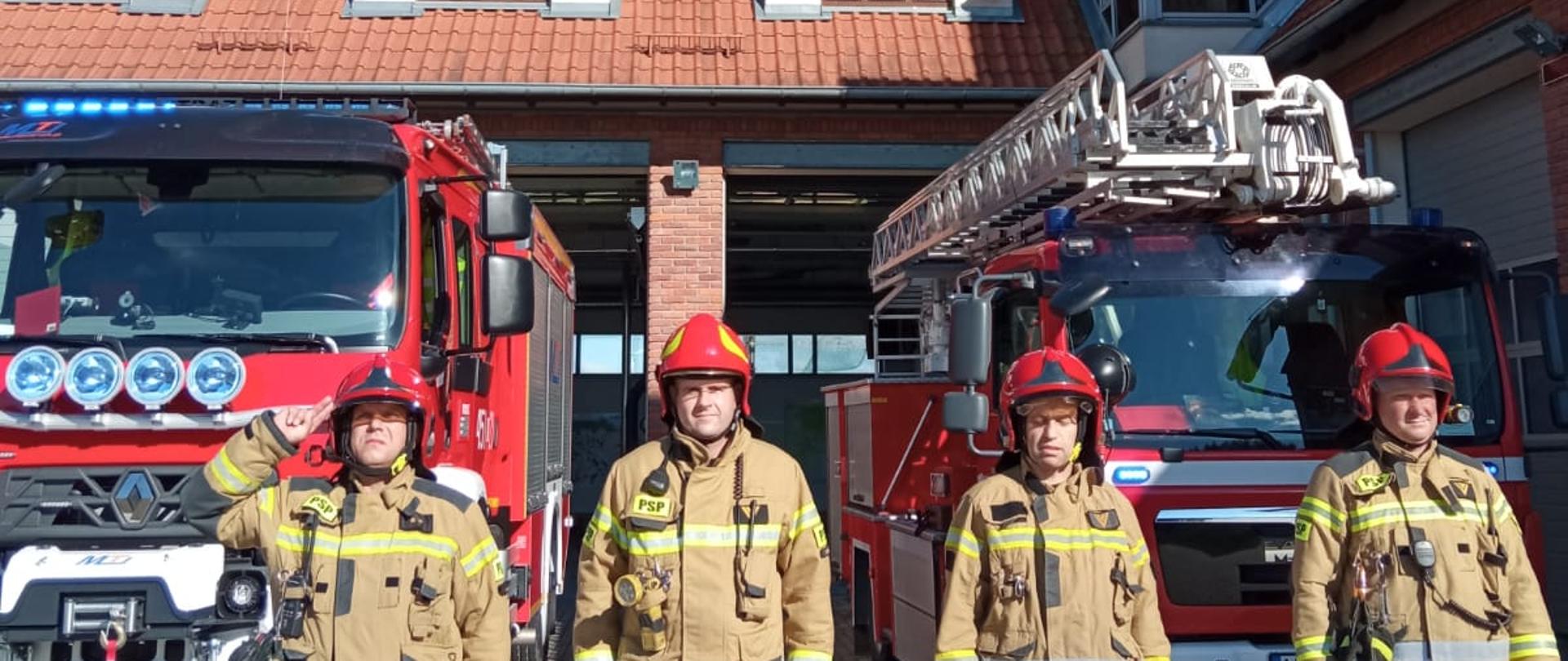 Zdjęcie przedstawia czterech strażaków z Komendy Powiatowej Państwowej Straży Pożarnej w Gołdapi, którzy uczcili 78. rocznicę wybuchu Powstania Warszawskiego poprzez wyjazd pojazdów pożarniczych przed garaże włączając o godz. 17:00 sygnały świetlne i dźwiękowe i oddając honor. W tle widać budynek strażnicy
