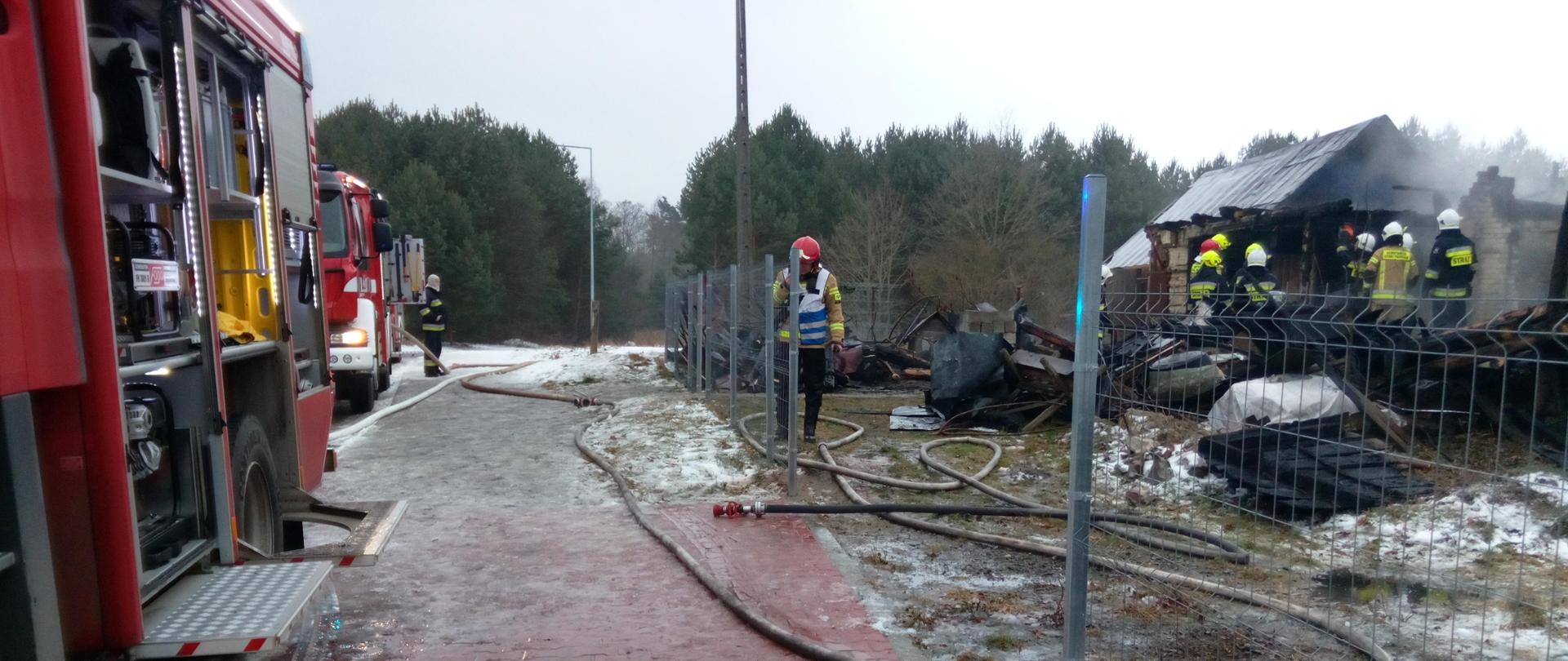Na zdjęciu widzimy: po prawej stronie drewniany budynek, który uległ spaleniu, po lewej samochody pożarnicze biorące udział w akcji ratowniczo-gaśniczej. Widać silne zadymienie nad budynkiem oraz pracujących ratowników.