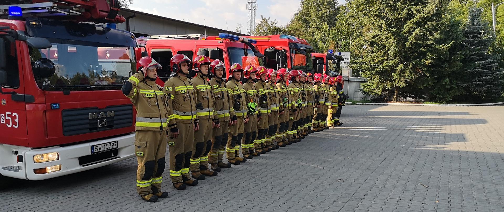 Strażacy oddają hołd, obok stoją samochody pożarnicze.