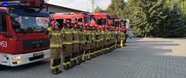 Strażacy oddają hołd, obok stoją samochody pożarnicze
