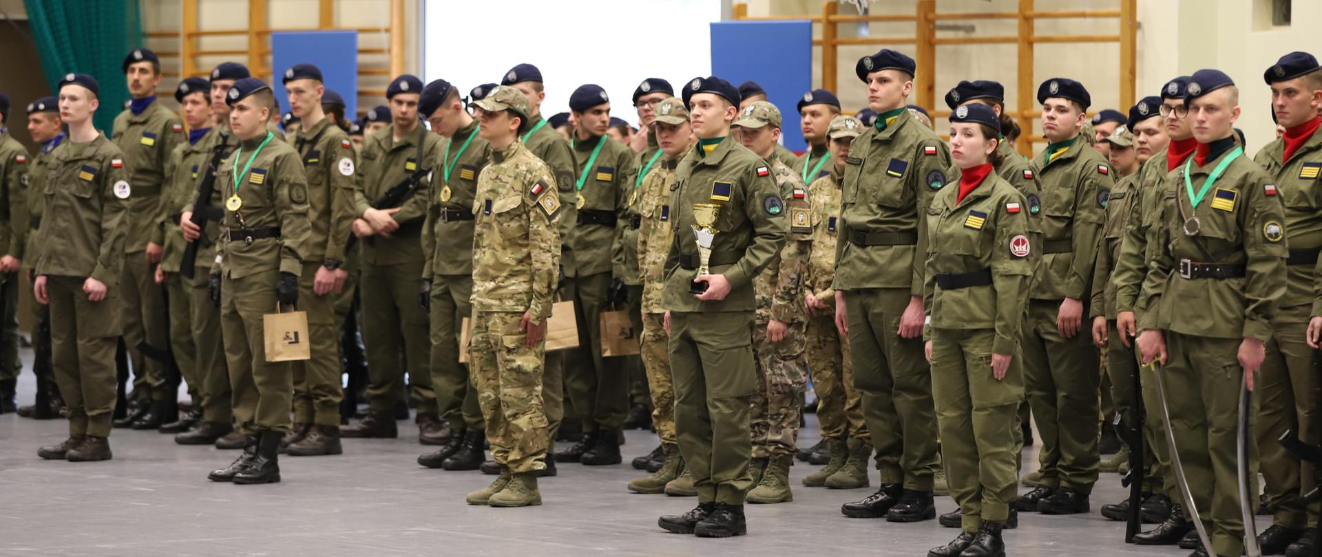 Grupa uczniów klas mundurowych stoi w hali sportowej podczas uroczystości. Część uczestników ma medale na szyjach, jedna osoba trzyma puchar. W tle widoczny kosz do koszykówki i banery sponsorów.