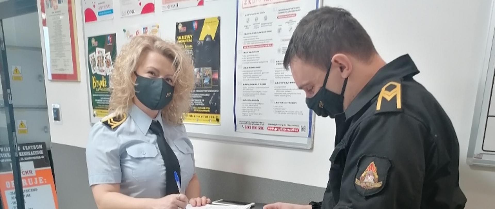 Wnętrze Powiatowego Centrum kulturalno-Rekreacyjnego we Włoszczowie, kobieta i mężczyzna w mundurach strażackich stoją przy stoliku i uzupełniają ankiety do rejestracji krwiodawców. 