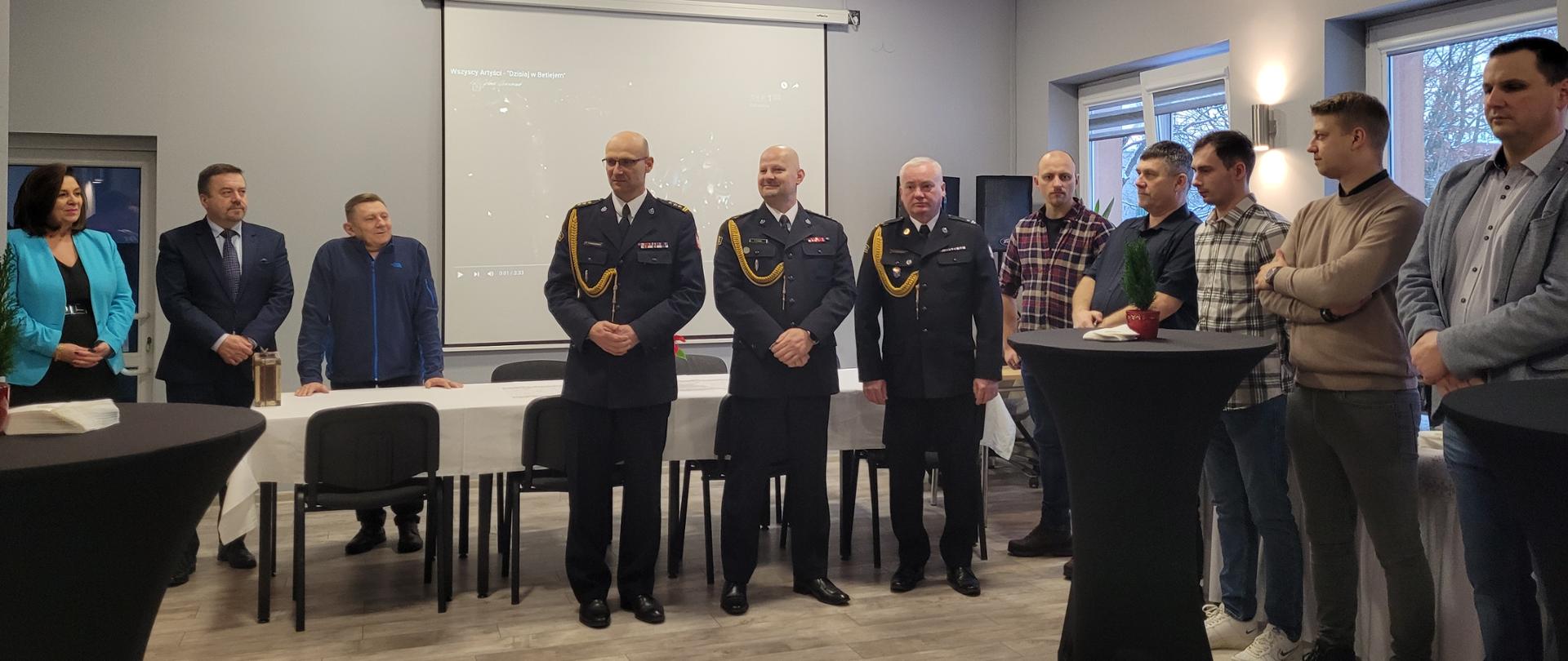 Na środku sali wykładowej stoją kolejno od lewej strony komendant powiatowy PSP w Żyrardowie st. bryg. Mariusz Tymoszewicz, zastępca Mazowieckiego komendanta wojewódzkiego PSP st. bryg. Tomasz Cybul, zastępca komendanta powiatowego PSP w Żyrardowie mł. bryg. Marek Krawczyk. Po ich prawej stronie stoją przedstawiciele władz samorządowych, a po lewej strażacy w ubraniach cywilnych.
