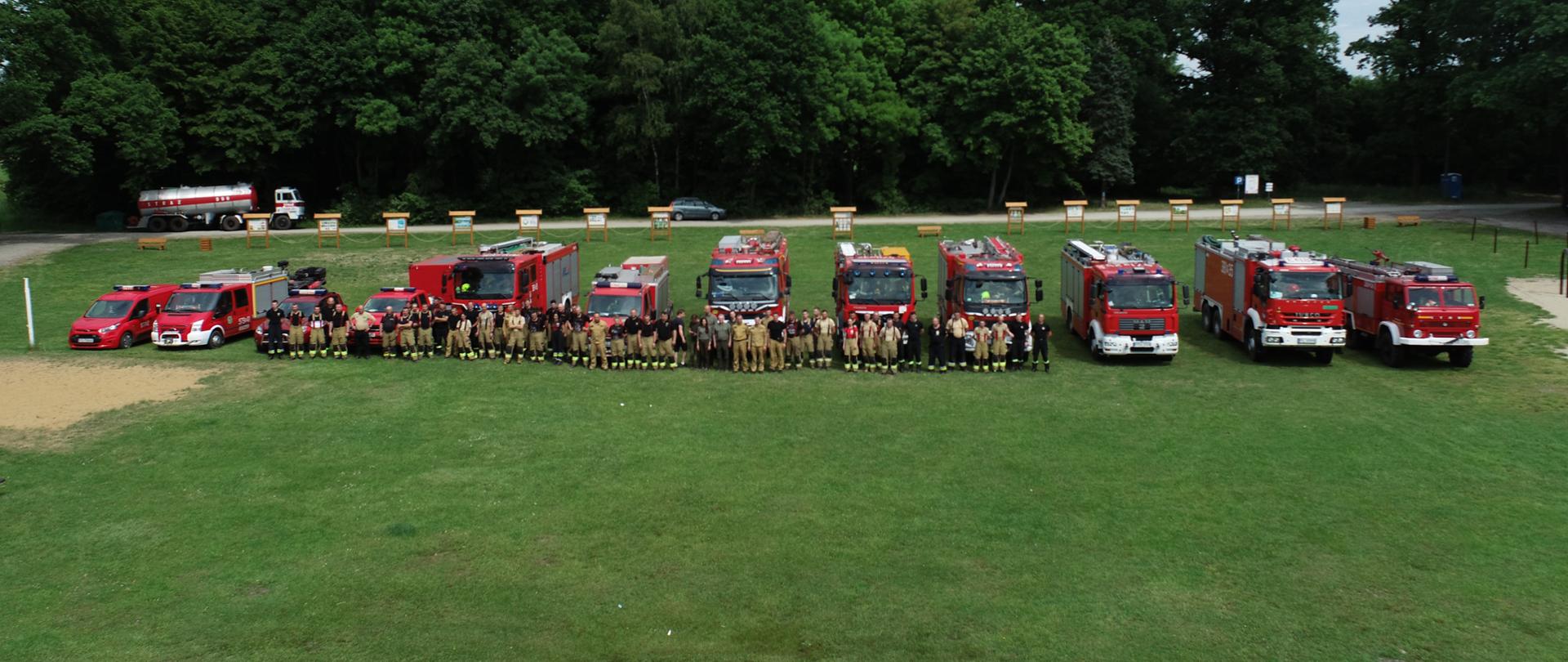 Ćwiczenia WOO. Teren Leśnictwa Górzno. Na terenie rekreacyjnym, na tle lasu stoją wszystkie pojazdy pożarnicze oraz strażacy uczestniczący w ćwiczeniach. Uczestnicy pozują do wspólnego zdjęcia.