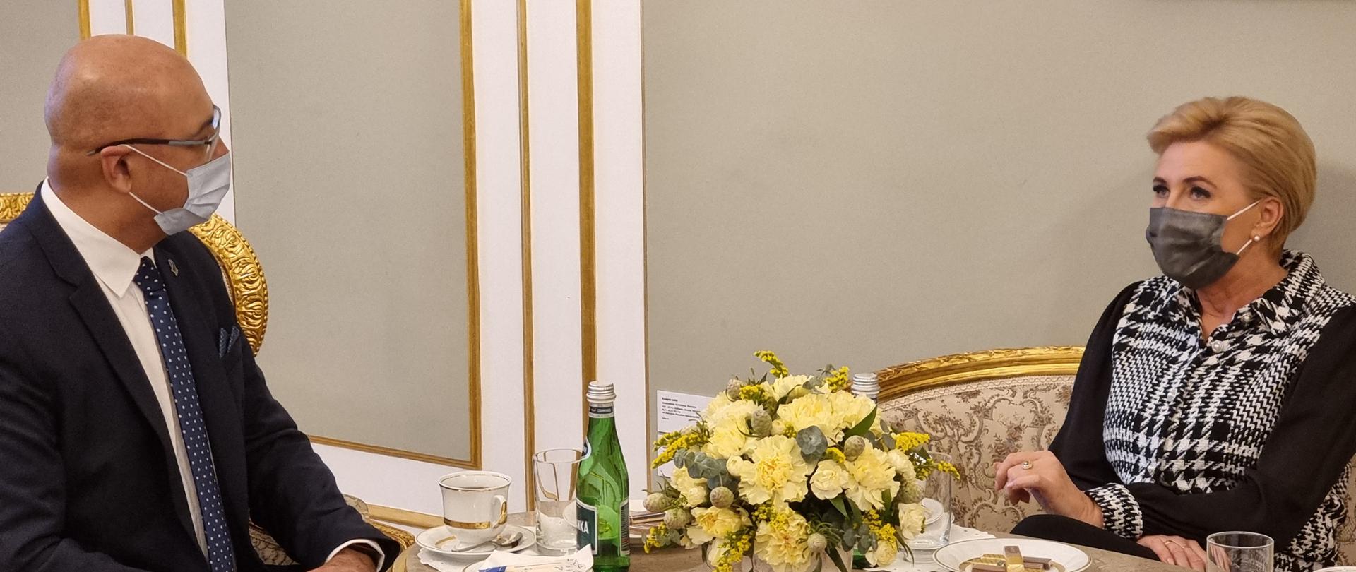 Główny Inspektor Transportu Drogowego Alvin Gajadhur wraz z Małżonką Prezydenta RP Agatą Kornhauser-Dudą siedzą przy zabytkowym stoliku, na którym stoi wazon z kwiatami. Główny Inspektor trzyma złożone dłonie na kolanach i z uwagą słucha słów Pierwszej Damy.