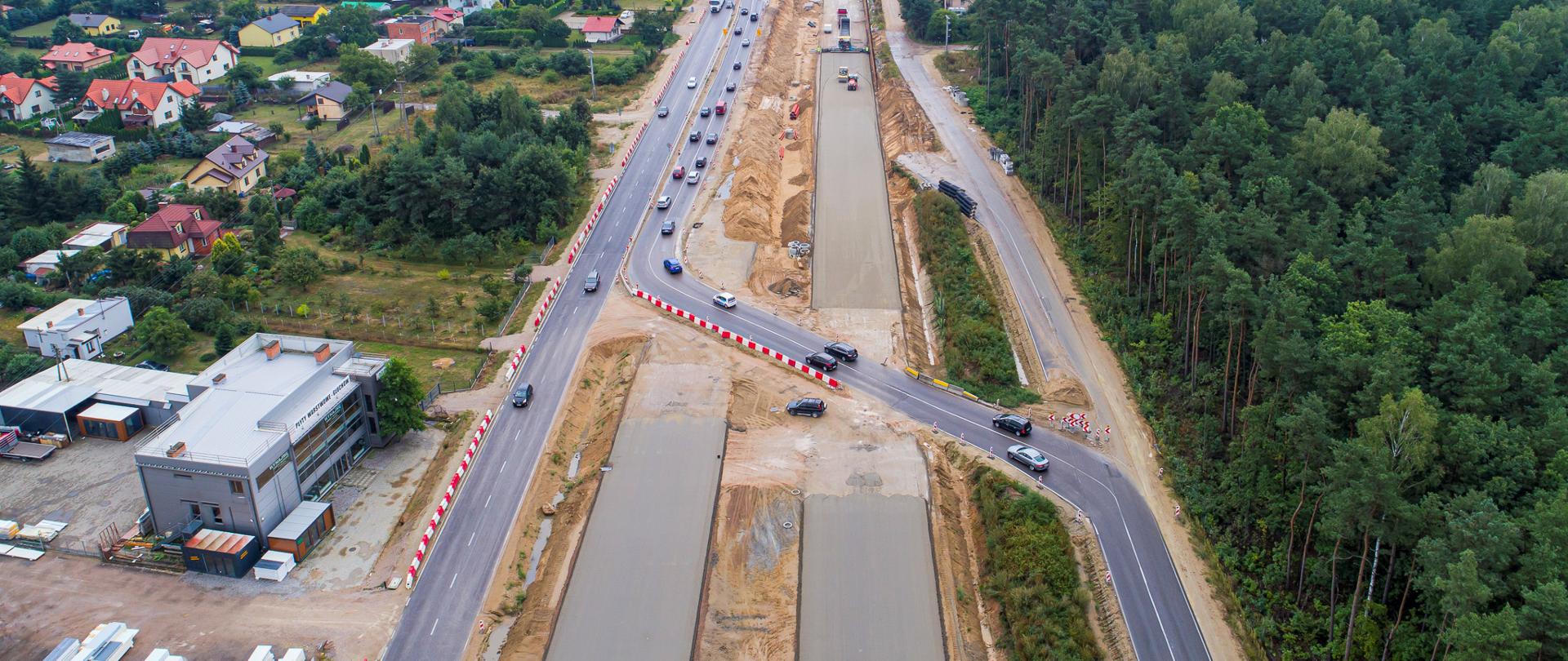 Widok z lotu ptaka na budowę drogi. Widać ślad nowej trasy oraz zaprojektowane objazdy dla bieżącego ruchu. Po lewej stronie widoczne zabudowania, a po prawej drzewa.