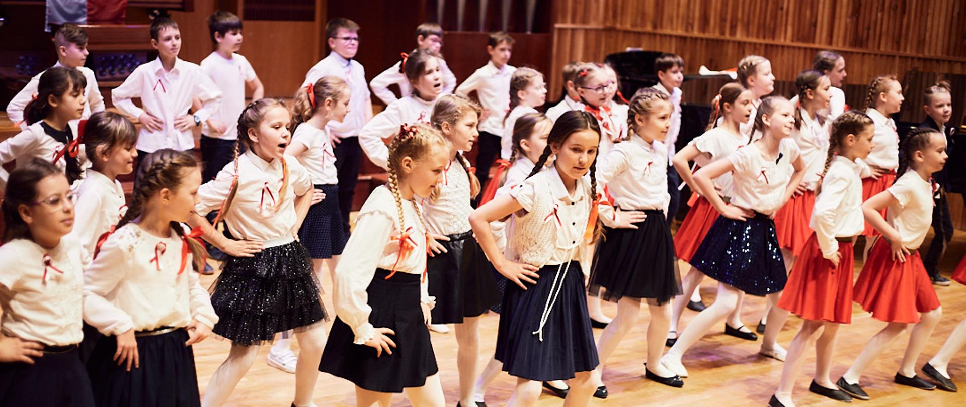 Na zdjęciu dzieci ubrane na galowo, w trzech szeregach. tańczą.