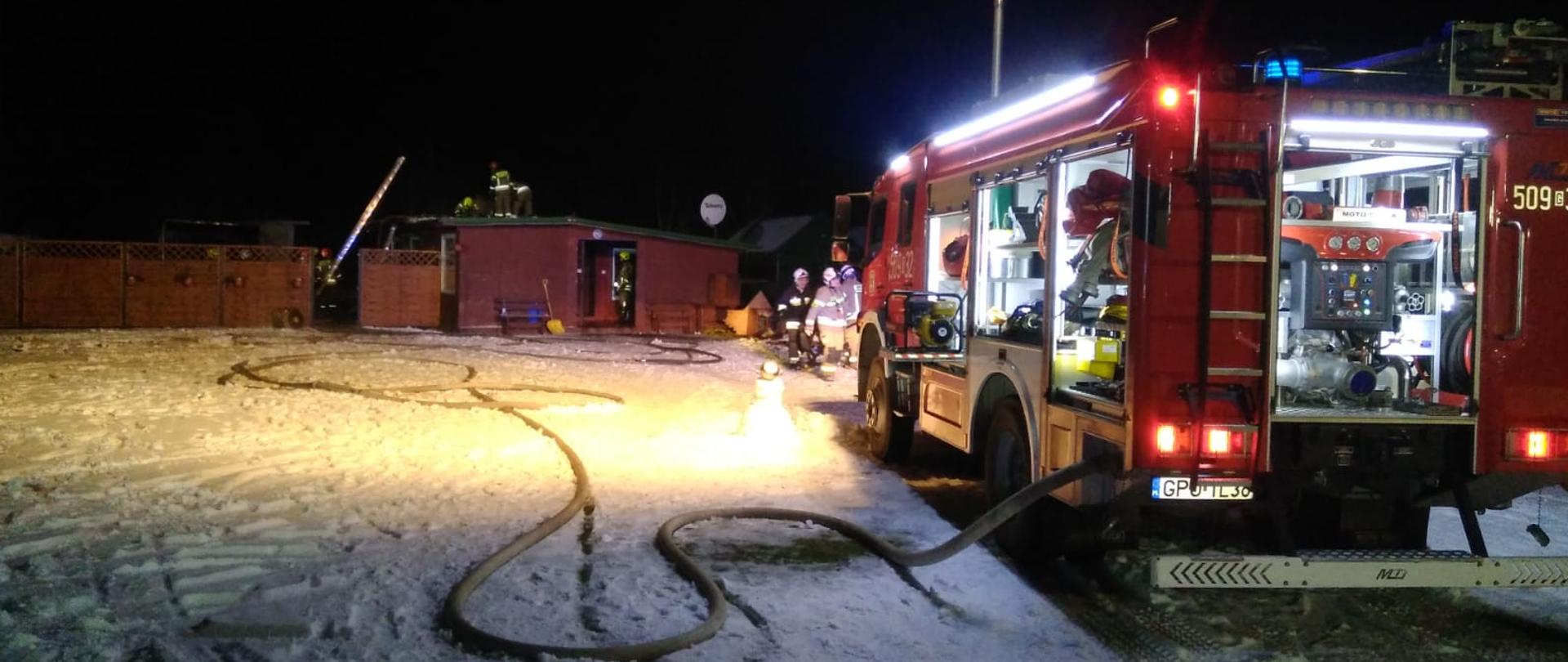 Na zdjęciu widać w porze nocnej budynek mieszkalny o raz samochód straży pożarnej z włączoną lampą oświetlającą teren akcji ratowniczej.