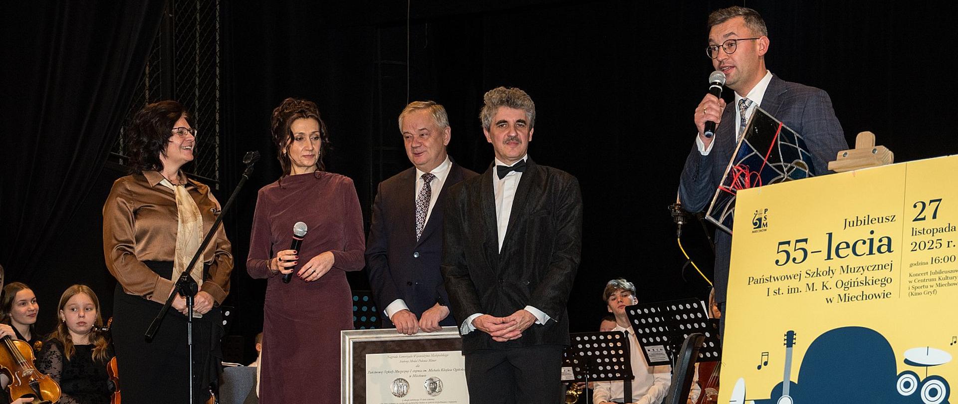 Grupa dorosłych na scenie podczas uroczystości jubileuszowej szkoły muzycznej; jedna osoba przemawia do mikrofonu, inni trzymają oprawiony dokument, obok stoi duży plakat wydarzenia.