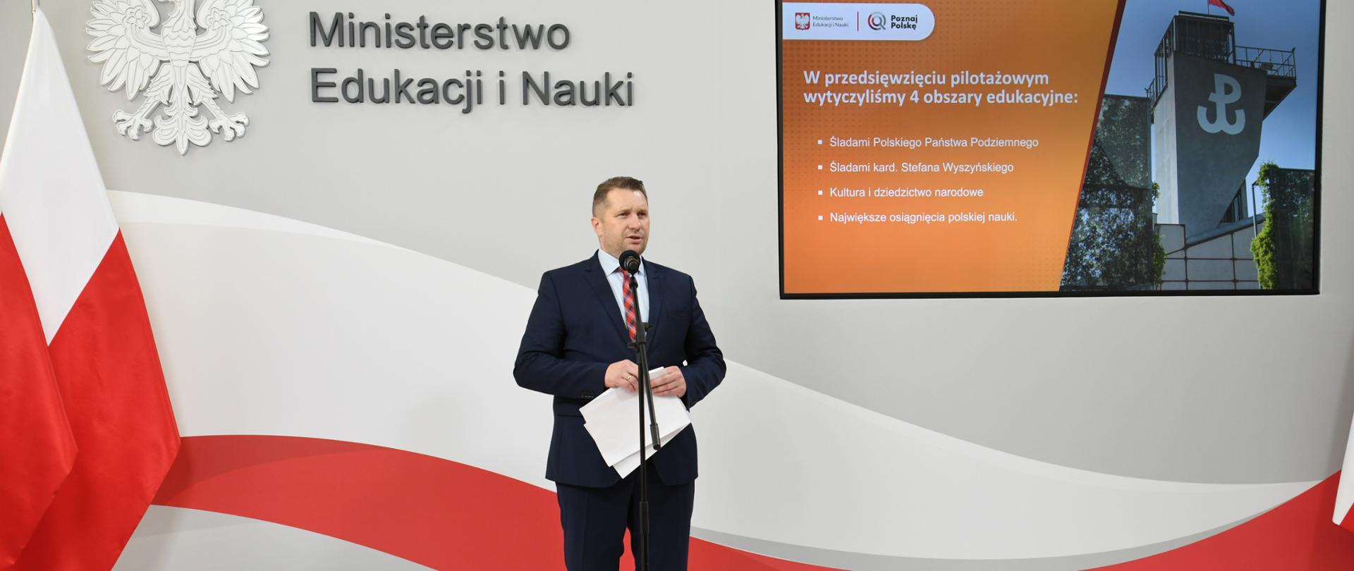 Minister Przemysław Czarnek stoi przy mikrofonie. W ręku trzyma dokumenty. W tle z lewej strony ekran ze zdjęciem muzeum powstania warszawskiego oraz napisem 