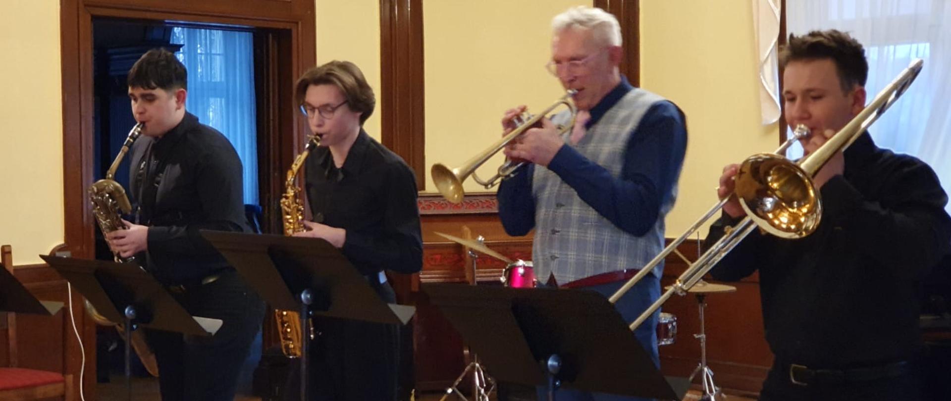 Zdjęcie kolorowe. Czterech muzyków w strojach galowych na scenie - grają na saksofonach, trąbce i puzonie. W tle zestaw perkusyjny.